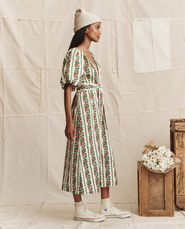 A woman in THE GREAT. PS26 The Fairground Dress – Tapestry Floral, with a belted waist, knit hat, and white sneakers, stands sideways near crates of white flowers against a fabric backdrop.