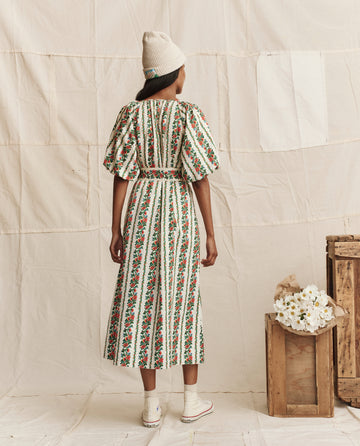 A woman faces away, wearing THE GREAT. PS26 The Fairground Dress in Tapestry Floral with a white knit hat and sneakers, near wooden crates and white flowers against a neutral fabric backdrop.