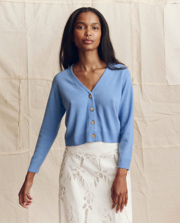 A woman stands in front of a beige fabric backdrop wearing THE GREAT. PS26 The Cashmere Tiny V Neck Cardigan in Soft Blue and a white lace skirt.