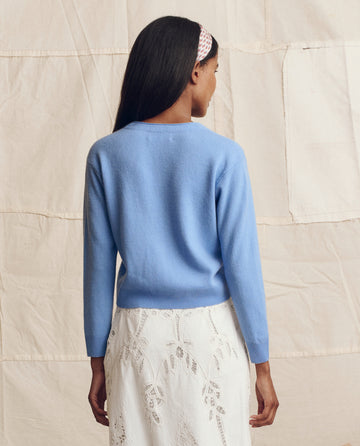 A woman with long dark hair wears THE GREAT. PS26 The Cashmere Tiny V Neck Cardigan in Soft Blue and a white lace skirt, standing with her back to a neutral textured background.