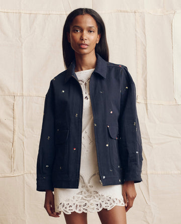 A woman stands against a neutral backdrop, wearing THE GREAT. PS26 The Folsom Jacket—navy cotton twill with tossed floral embroidery—styled over a white lace dress.
