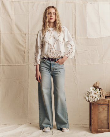 A person stands indoors wearing THE GREAT. PS26 The Picnic Top in cream, light blue wide-leg jeans, and white sneakers. A crate filled with white flowers sits to the right.