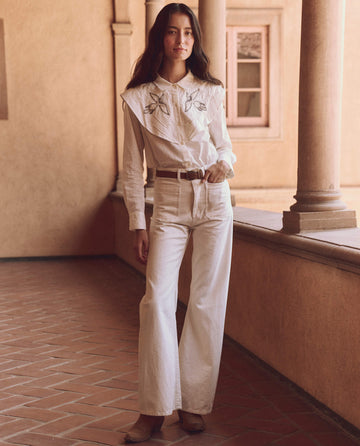 A woman in a white western-style outfit, featuring THE GREAT. SP25 D1's The Beaded Partridge Top in cream, stands in a tiled corridor with columns, her arms relaxed by her sides.