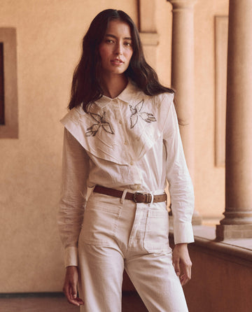 A person with long hair wearing THE GREAT. SP25 D1's The Beaded Partridge Top in cream and white pants stands gracefully near a corridor lined with columns.
