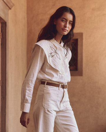 Indoors, a person dons THE GREAT.'s The Beaded Partridge Top in cream, paired with white pants, exuding elegance with a neutral expression.