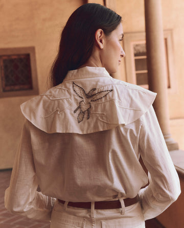 In a courtyard with classical columns, a woman in white wears THE GREAT. SP25 D1's Cream Beaded Partridge Top, showcasing intricate bird embroidery on the back.