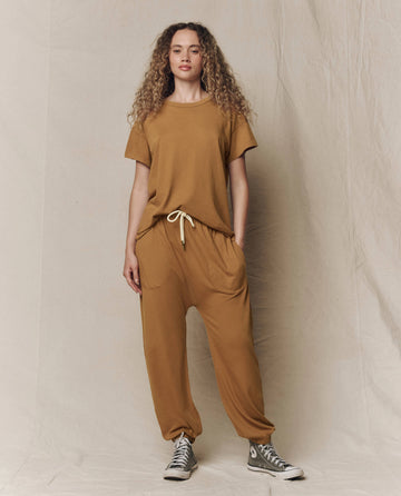 A person wearing THE GREAT.'s The Jersey Jogger Pant in Cork with a loose matching brown T-shirt and gray high-top sneakers stands against a beige fabric backdrop.