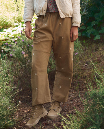 A person stands outdoors in THE GREAT. SP26 TGO The Embroidered Outpost Pant—Khaki With Cream Daisies—tan boots, and a light jacket, surrounded by lush greenery.