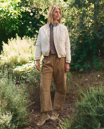 Standing outdoors in a garden, a person wears a white quilted jacket, patterned shirt, tan lace-up boots, and THE GREAT. SP26 TGO The Embroidered Outpost Pant in khaki with cream daisies.