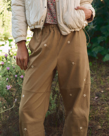 Person wearing THE GREAT. SP26 TGO's The Embroidered Outpost Pant in khaki with cream daisies, standing outdoors near greenery and pink flowers.
