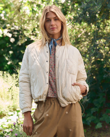 A woman stands outdoors among green foliage wearing THE GREAT. SP26 TGO's Reversible Ultra Light Puffer in Khaki Bandana W/ Cream over a floral shirt, tan pants with small white flowers, and a blue neck scarf.
