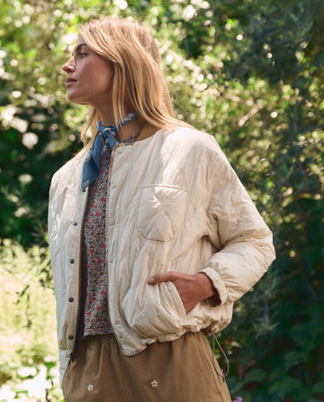 Outdoors, a person wears THE GREAT. SP26 TGO The Reversible Ultra Light Puffer in Khaki Bandana W/ Cream over a floral top, paired with beige floral pants and a blue scarf, surrounded by greenery.