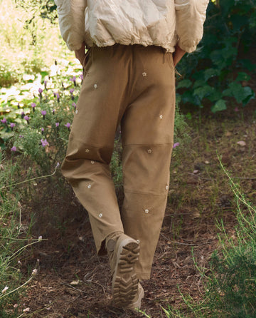 A person stands on a dirt path in THE GREAT. SP26 TGO's Embroidered Outpost Pant—Khaki With Cream Daisies—paired with beige shoes and a light jacket. Lush greenery and flowers form the backdrop.