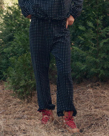 Person in The Campfire Sweatpant. -- Black Dot by THE GREAT. F26 TGO and red hiking boots stands on dry grass with green bushes behind them.