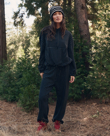 A woman stands among tall trees, wearing THE GREAT. F26 TGO The Campfire Sweatpant. -- Black Dot, a matching knit hat, and red boots in a lush forest setting.