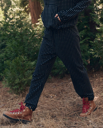 A person in THE GREAT. F26 TGO The Campfire Sweatpant. -- Black Dot and brown hiking boots with red laces walks on a pine needle-covered forest floor.