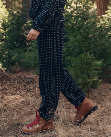 A person in The Campfire Sweatpant. -- Black Dot by THE GREAT. F26 TGO walks through a forest with dry grass and green trees in the background, wearing tan hiking boots.