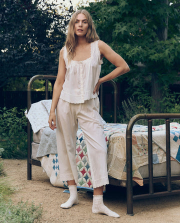 A woman stands outdoors by a metal-framed bed draped in quilts, wearing THE GREAT. CORE SLEEP The Eyelet Easy Sleep Pant in True White, with lush greenery in the background.