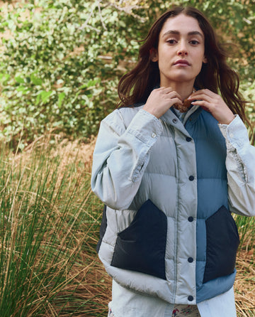 Outdoors amid green foliage, a person adjusts a scarf while wearing THE GREAT. F25 TGO's Polar Vest – Navy Colorblock with PrimaLoft insulation over a button-up shirt.