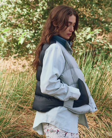 A person with long brown hair stands in tall grass, wearing a light blue shirt and THE GREAT. F25 TGO’s The Polar Vest in Navy Colorblock, crafted from recycled materials with PrimaLoft insulation for warmth.