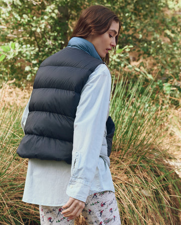 A person wearing THE GREAT. F25 TGO The Polar Vest in Navy Colorblock, a light blue shirt, and patterned pants stands outdoors among tall grass and lush greenery.