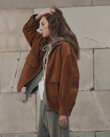 Wearing THE GREAT. F26 D1 The Suede Folsom Jacket in Cognac with an oversized fit, a gray hoodie, and sweatpants, the person stands sideways in front of a light stone wall, touching their head.