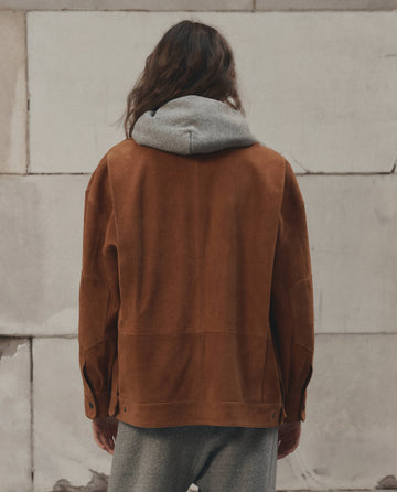 A person with long hair is seen from behind, wearing THE GREAT. F26 D1′s The Suede Folsom Jacket in Cognac—an oversized brown suede style—layered over a gray hooded sweatshirt, standing before a textured wall background.
