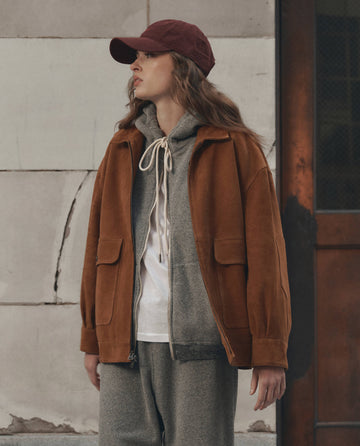 A person wearing THE GREAT. F26 D1 The Suede Folsom Jacket in Cognac, along with a gray hoodie, gray sweatpants, white shirt, and a maroon cap stands outdoors near a concrete wall and a door.