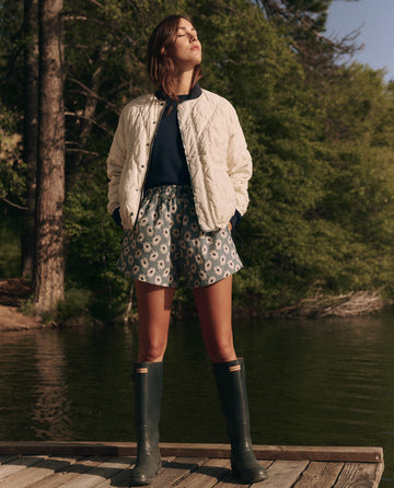 A person stands on a wooden dock by the lake, wearing THE GREAT. SP25 TGO's Reversible Ultra Light Down Puffer in Falling Daisy and Cream over patterned shorts with green boots, surrounded by serene trees.