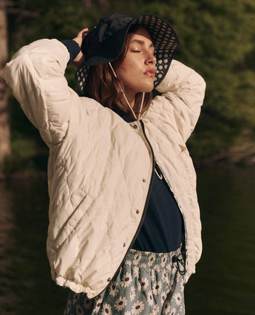 A person stands outdoors, eyes closed and hands raised, wearing THE GREAT. SP25 TGO's The Reversible Ultra Light Down Puffer in Falling Daisy and Cream with floral pants and a wide-brimmed hat.