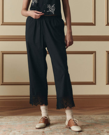 A person stands indoors wearing The Rosette Crochet Pant in black by THE GREAT. SP26 D2, paired with a black top featuring white embroidery, cream socks, and two-tone oxford shoes.