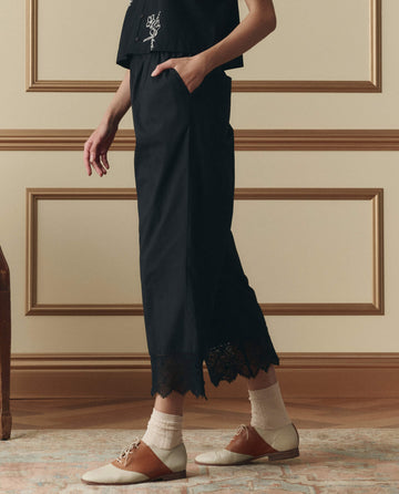 A person stands indoors wearing THE GREAT. SP26 D2 The Rosette Crochet Pant in black, featuring lace-trimmed wide legs, cream socks, and brown-and-cream lace-up shoes; only the lower half is visible.