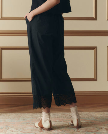 Wearing THE GREAT.'s The Rosette Crochet Pant in black, paired with white socks and two-tone oxford shoes, a person stands on a patterned rug before a paneled wall.