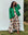 A person stands on grass wearing a green quilted jacket over The Eden Dress in Oasis Floral by THE GREAT. PS25, with brown boots against a plain backdrop.