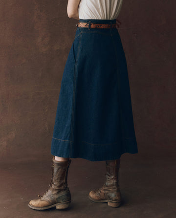 A person stands against a brown background wearing a white top, THE GREAT. F25 D1 The Field Skirt in Midnight Rinse, a brown belt, and worn brown lace-up boots. Their face is not visible.