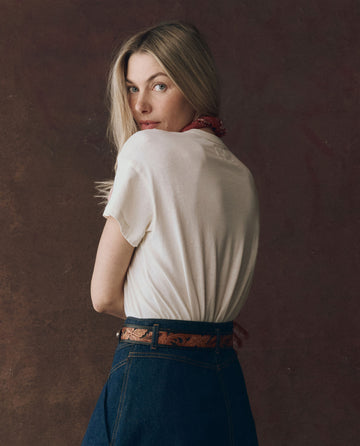 A person with long blonde hair wears THE GREAT. F25 D1’s The Boxy Crew, Graphic tee in Washed White with Rodeo Star Graphic, along with a red bandana and blue jeans, looking over their shoulder against a dark background.