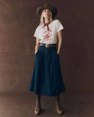 A person stands against a brown backdrop with hands in pockets, wearing a cowboy hat, white graphic tee, red bandana, lace-up boots, and THE GREAT. F25 D1 The Field Skirt in Midnight Rinse.