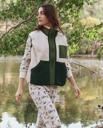 A woman stands by a pond in THE GREAT. F25 TGO "Plush Tundra Vest" in Cream & Army, layered over a printed long-sleeve. Trees and water set a vintage camping gear vibe.