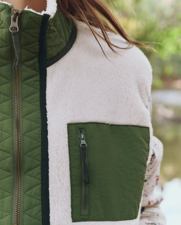 Close-up of someone wearing THE GREAT. F25 TGO Plush Tundra Vest—Cream & Army—a cozy fleece vest with green quilted details, zippered collar, and chest pocket inspired by vintage camping gear.