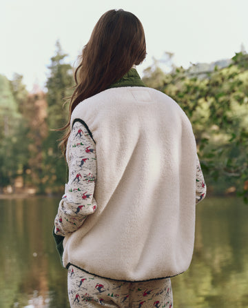 A person with long hair stands by a lake, wearing THE GREAT. F25 TGO's The Plush Tundra Vest in Cream & Army over a patterned outfit, facing away from the camera with trees in the background.