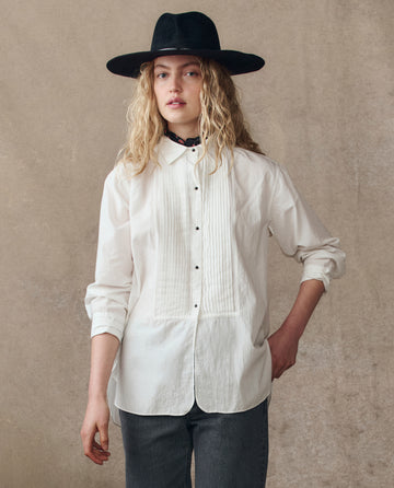 A person with long curly hair wearing THE GREAT. H25 COLLECTION's The Classic Tux Top in white, a wide-brimmed black hat, and dark pants stands against a neutral textured background.
