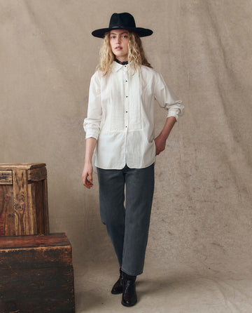 Wearing THE GREAT. H25 COLLECTION's The Classic Tux Top in white with dark jeans, black boots, and a wide-brimmed hat, a person stands indoors beside wooden crates against a neutral backdrop.