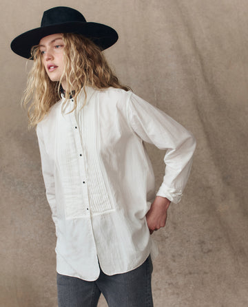 A person with long wavy hair wears THE GREAT. H25 COLLECTION's The Classic Tux Top.. in white, styled as a button-up shirt, paired with a wide-brimmed black hat and dark jeans, standing against a neutral textured background.