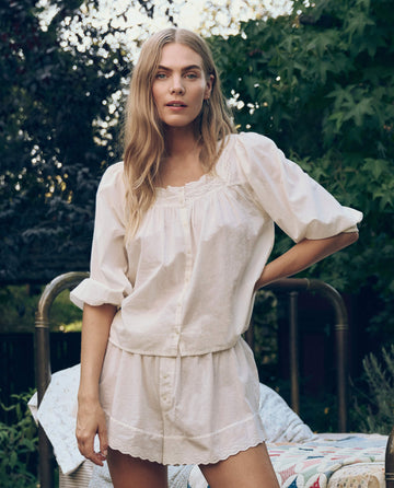 A woman with long blonde hair stands outdoors in greenery, wearing THE GREAT. CORE SLEEP's The Eyelet Button Sleep Top in True White—a loose, light-colored blouse with Victorian lace trim.