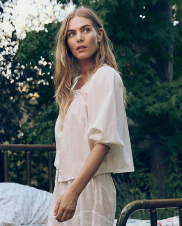 A woman with long blonde hair stands outdoors beside a metal bed frame, wearing THE GREAT. CORE SLEEP's Eyelet Button Sleep Top in True White, featuring delicate Victorian lace trim, with trees in the background.