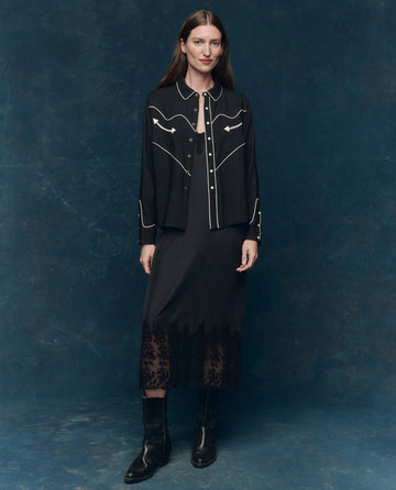 A woman stands against a blue textured backdrop, wearing THE GREAT. SP25 COLLECTORS' The Legacy Dress in black and black boots for a timeless fall look.