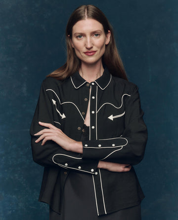 A woman with long brown hair stands with arms crossed against a dark blue background, wearing THE GREAT. F25 COLLECTORS The Western Shirt in black with cream detailing.
