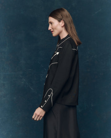 A woman with straight brown hair stands in profile against a dark blue background, wearing THE GREAT. F25 COLLECTORS The Western Shirt in Black with Cream and a skirt.
