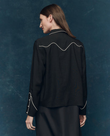 A woman faces away, wearing THE GREAT. F25 COLLECTORS The Western Shirt in black with cream details and pearl snaps, set against a dark blue textured background.