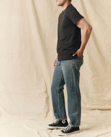 A person stands sideways against a beige fabric backdrop, wearing THE GREAT. FALL 23 MEN's Pure Knits Slim Tee in Solid -- Almost Black, paired with blue jeans and black high-top sneakers.